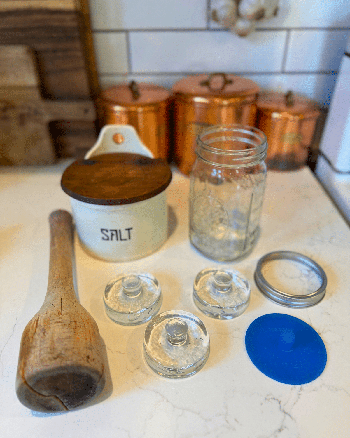 red sauerkraut fermenting tools wood pounder, salt, wide mouth mason jar, fermenting weights, rubber nippled lid and jar ring band on the counter