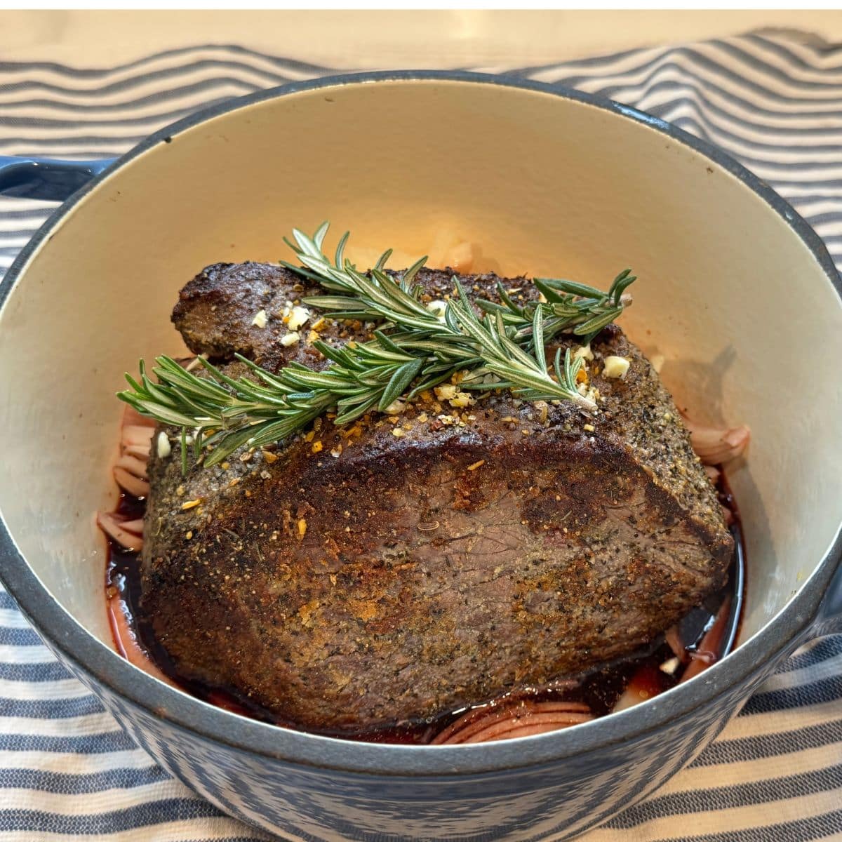 seasoned and seared rump roast in dutch oven with broth, onions and herbs on top