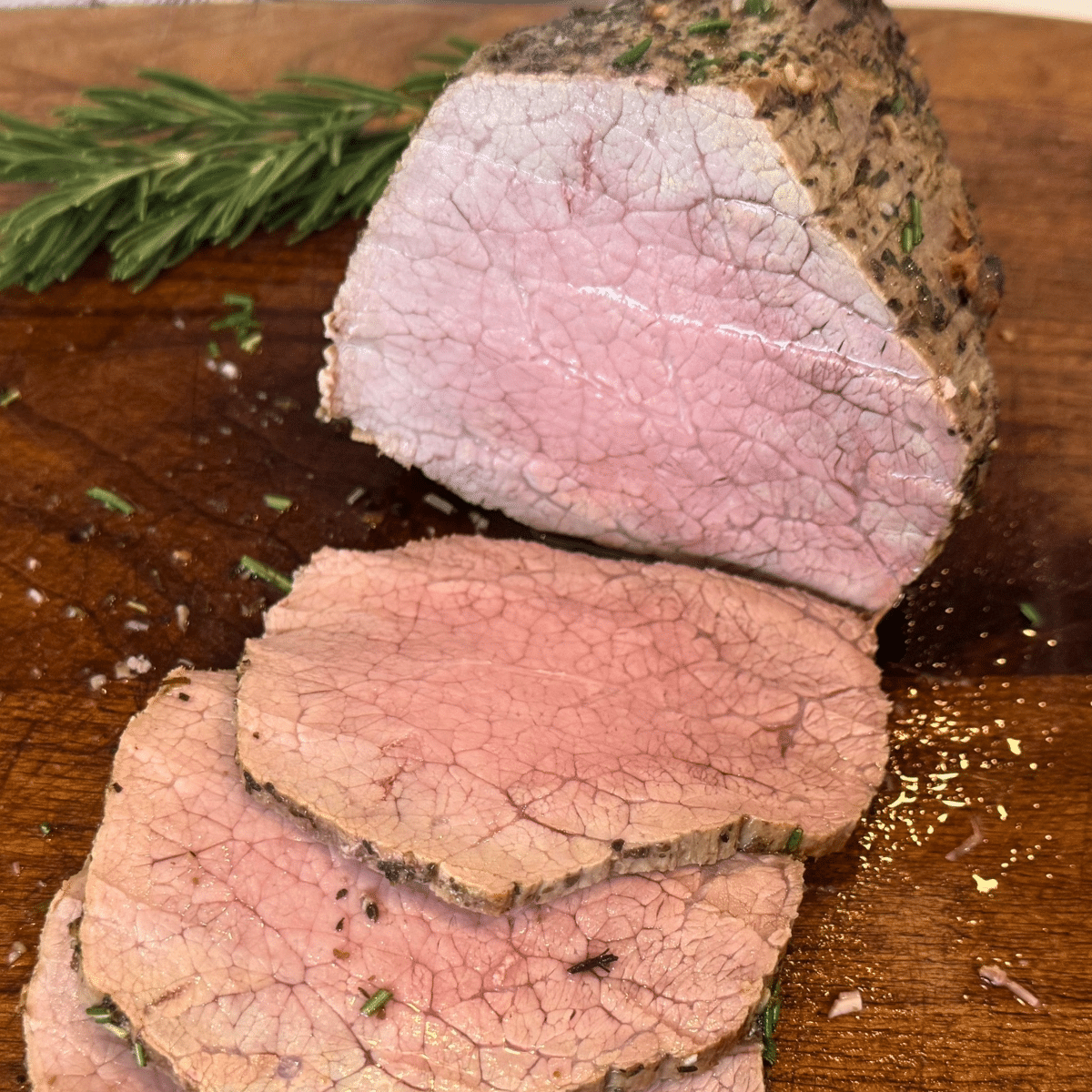 sliced perfectly done medium eye of round roast on wooden cutting board with a sprig of rosemary