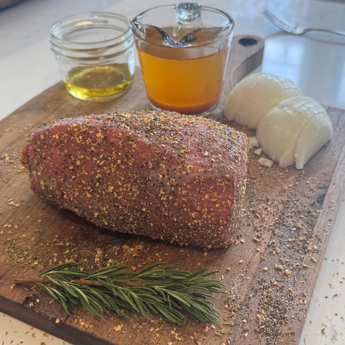 seasoned raw eye of round roast on cutting board with onions, broth, oil and rosemary