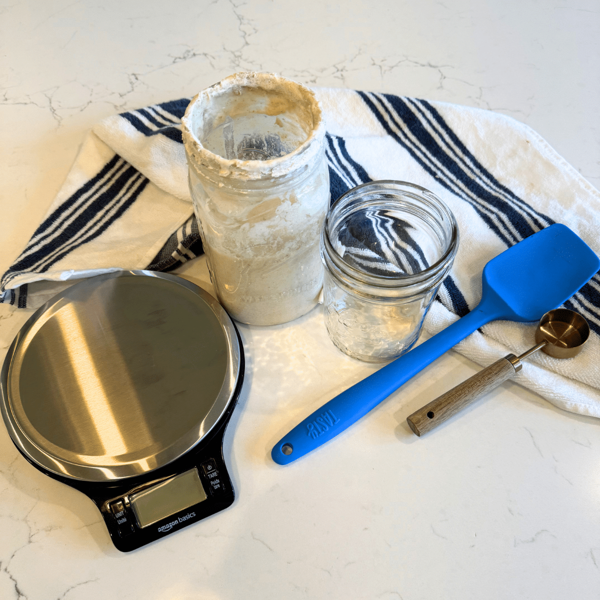 kitchen scale sourdough starter wide mouth mason jar rubber spatula and tablespoon on counter with a kitchen towel
