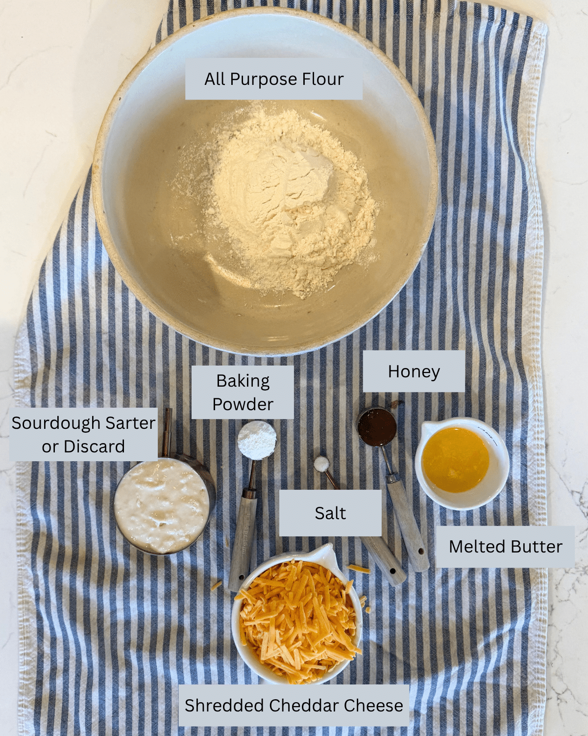 flour, sourdough starter, honey, butter, salt, baking powser, shredded cheddar cheese all labled and measured out on counter
