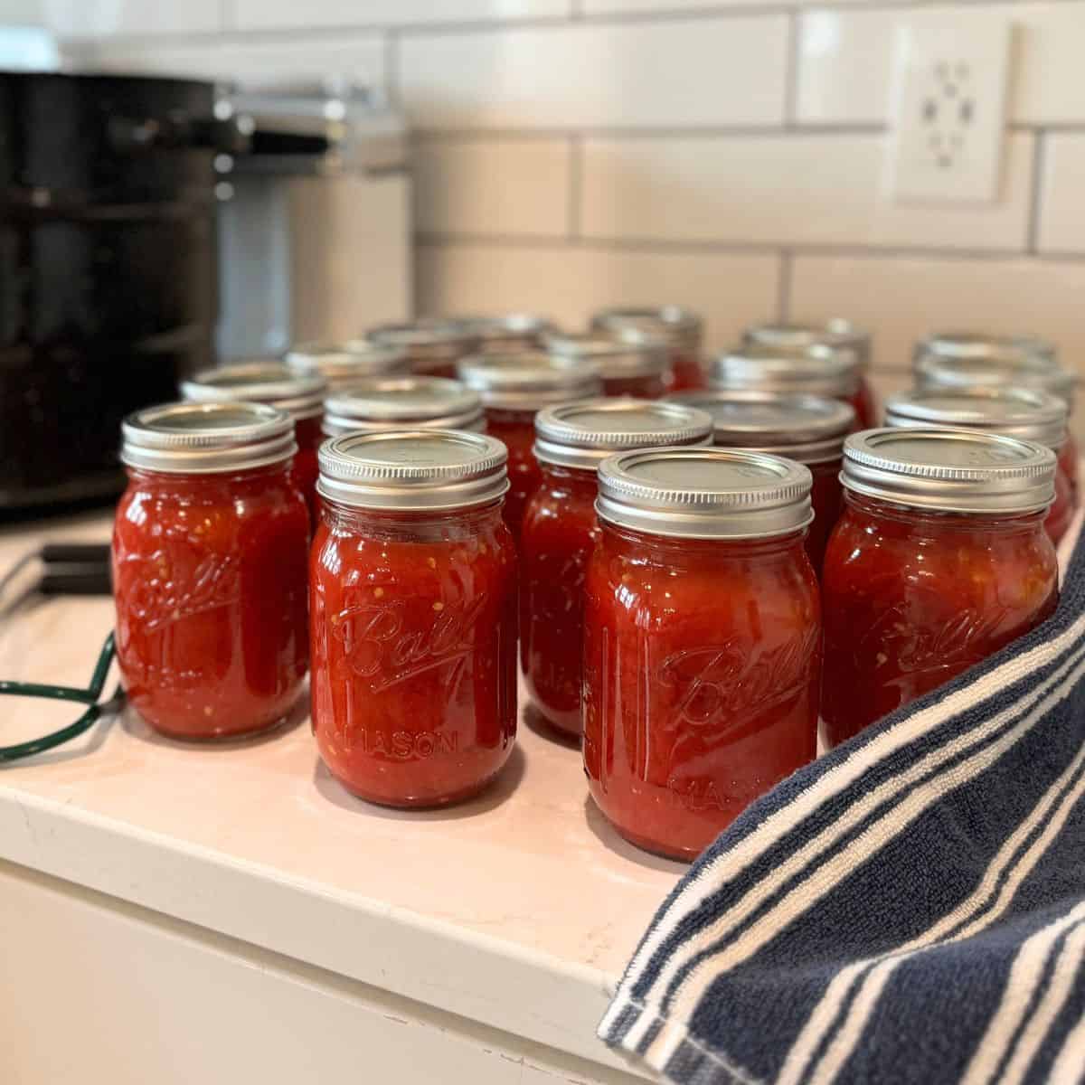 20 pints of home canned crushed tomatoes in jars on counter with water bath canner behind them.