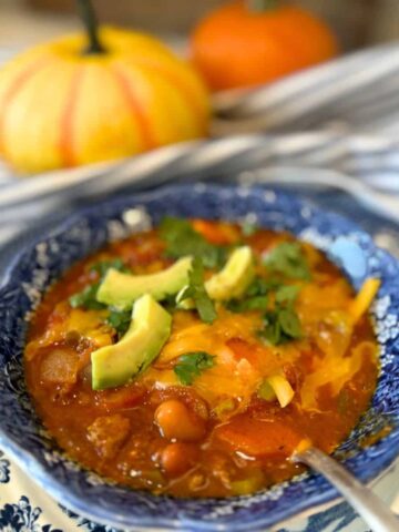 of blue bowl of pumpkin chili recipe garnished with avocado cheese and cilantro and small pumpkins in the background