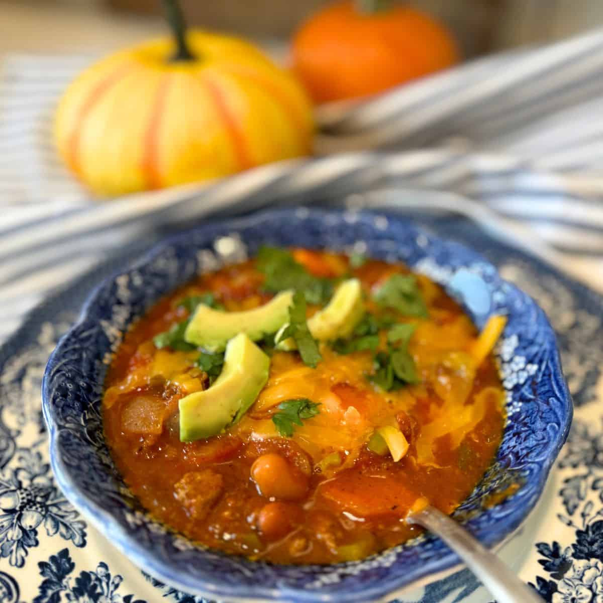 of blue bowl of pumpkin chili recipe garnished with avocado cheese and cilantro and small pumpkins in the background