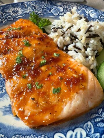 Baked Glazed Salmon served on a blue plate with chopped parsley, wild rice pilaf and cucumber slices.