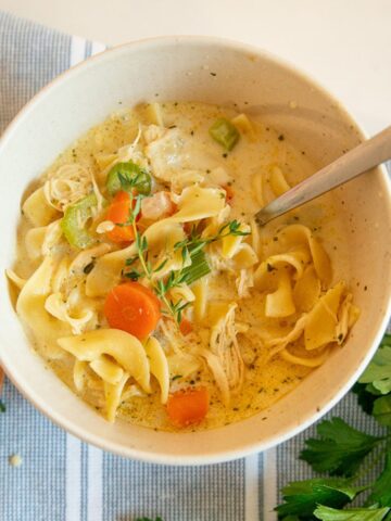 creamy chicken noodle soup in a bowl with thyme sprig, a dinner roll and parsley