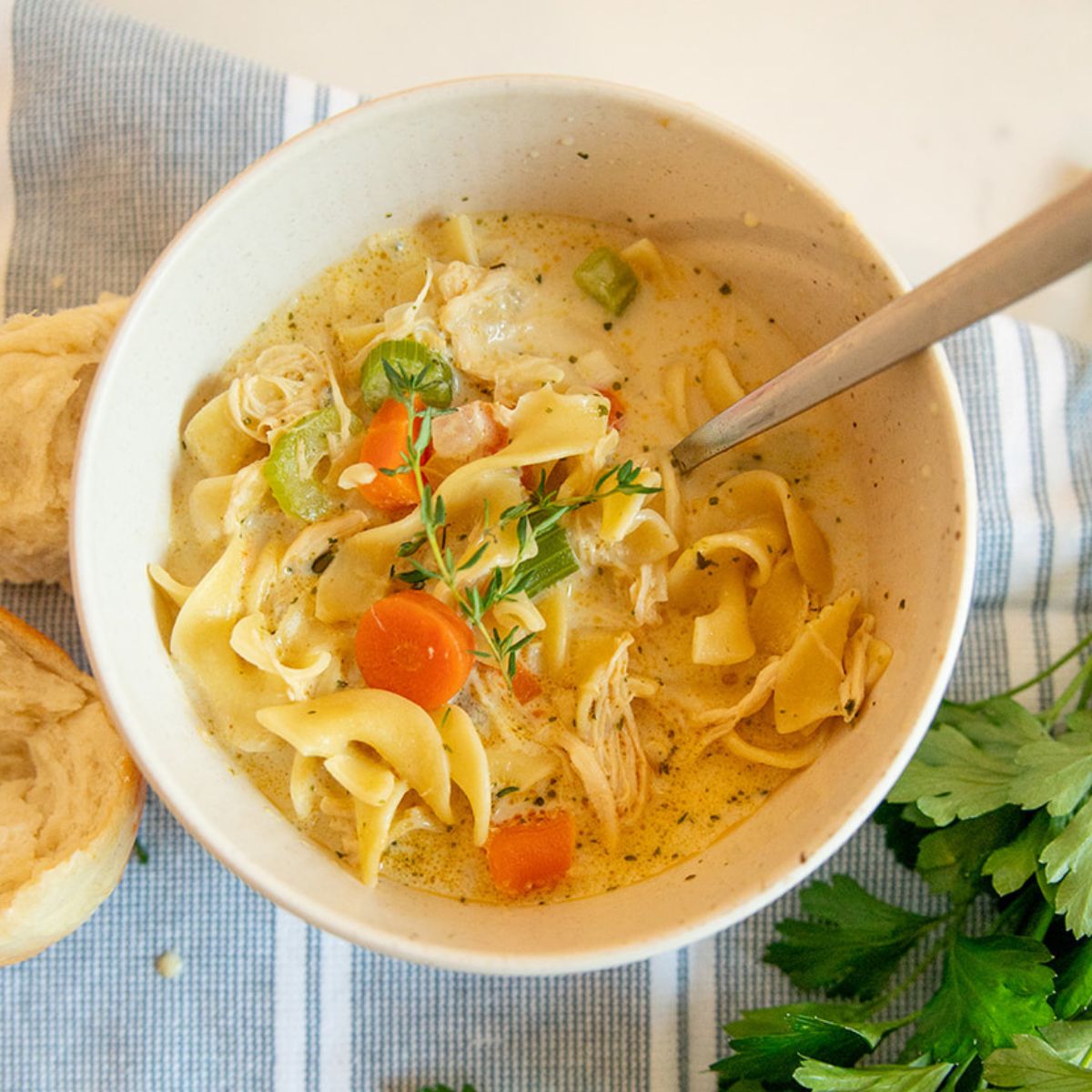 creamy chicken noodle soup in a bowl with thyme sprig, a dinner roll and parsley