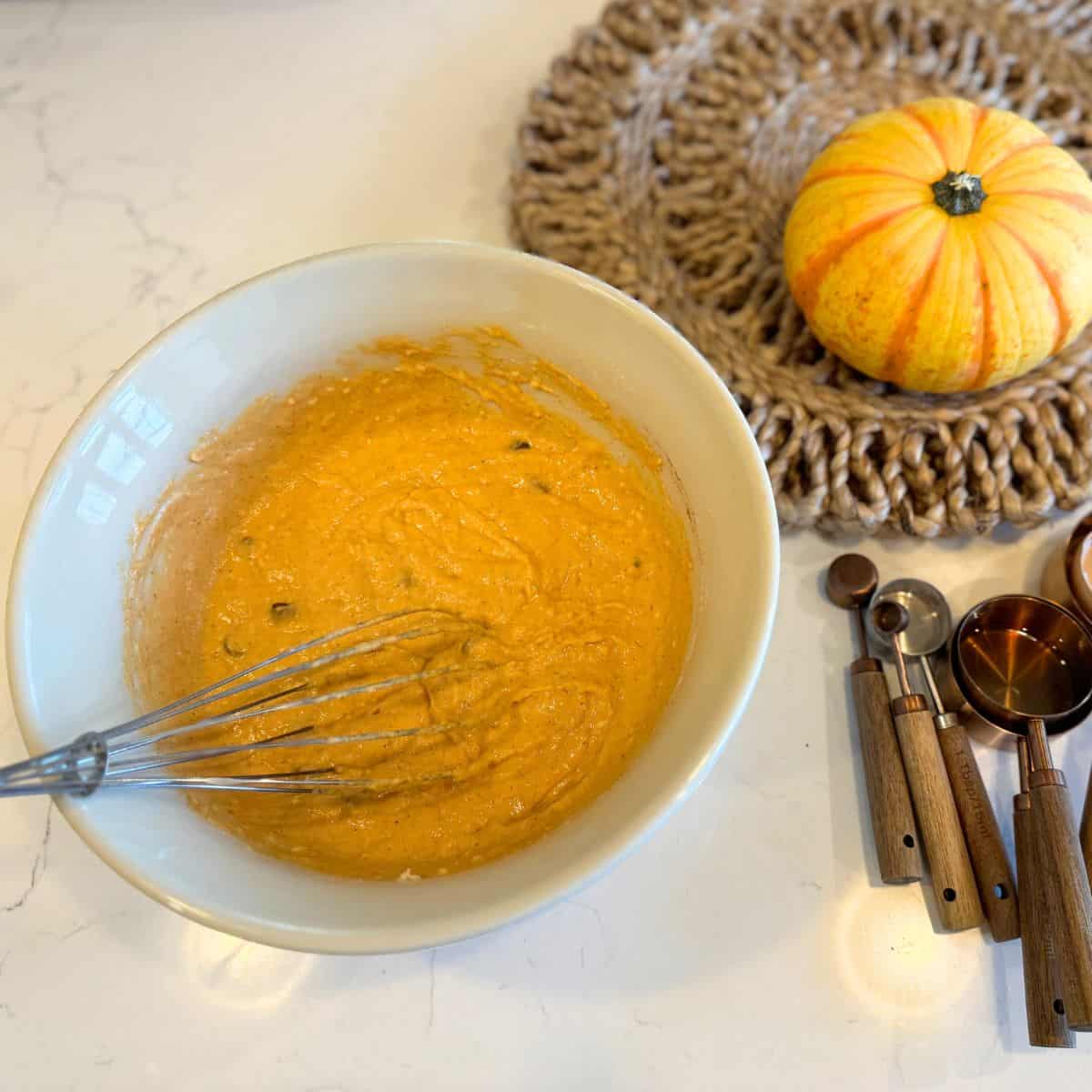 mixed pumpkin pancake batter in bowl with empty measuring cups, and a small pumpkin