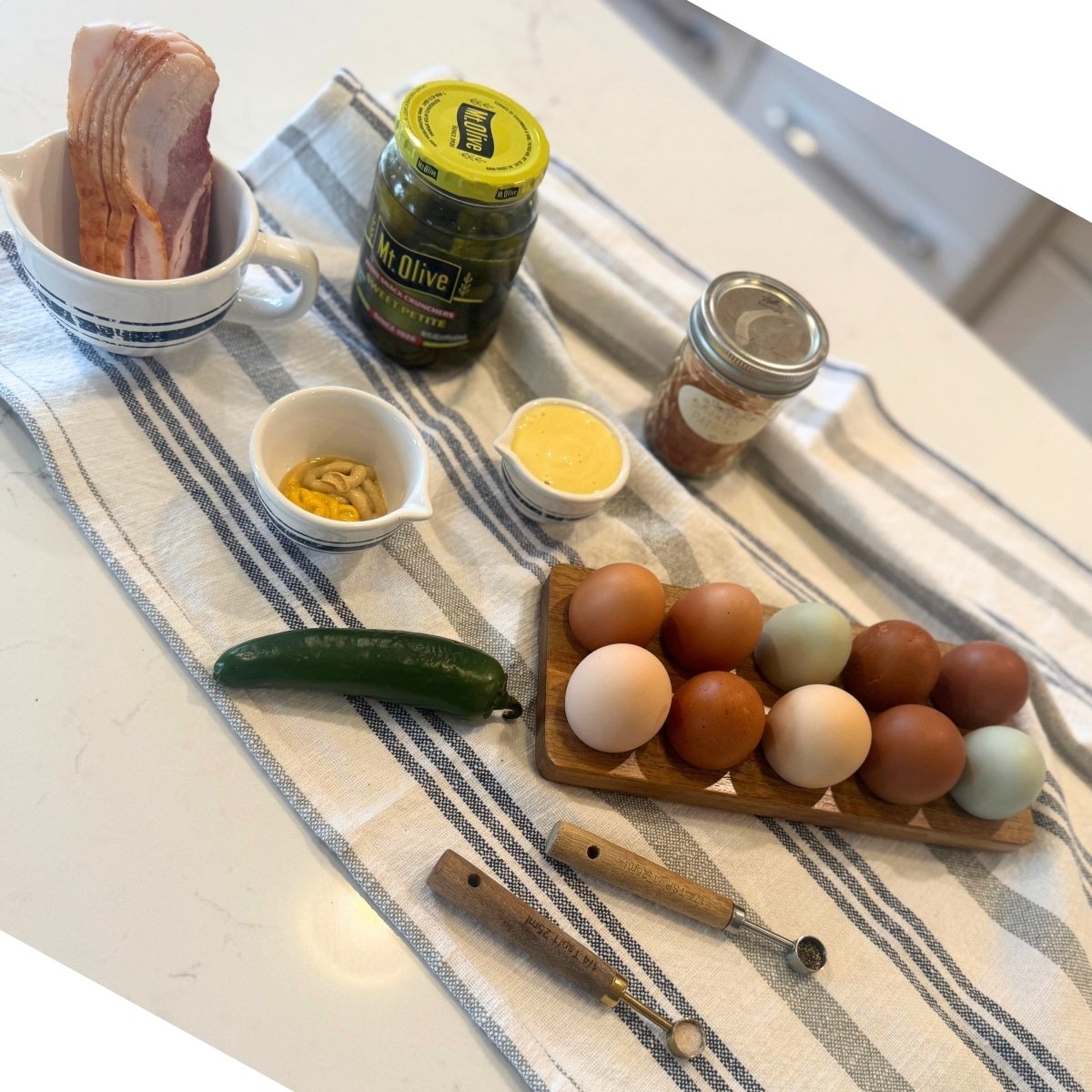 bacon jalapeno deviled eggs on counter: raw bacon, sweet pickled in a jar, smoked paprika in a jar, homemade mayo, yellow and dijon mustard in a dish, a jalapeno, salt, pepper and a dozen fresh eggs
