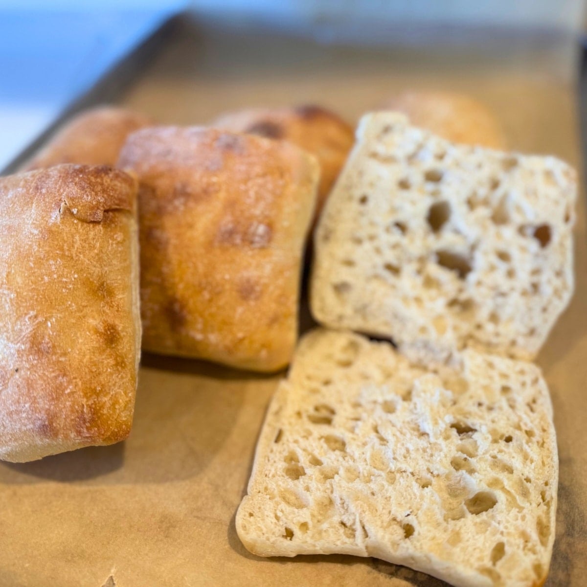 sousrdough ciabatta rolls onbaking sheet lined with parchment paper.. One open rolls to show texture