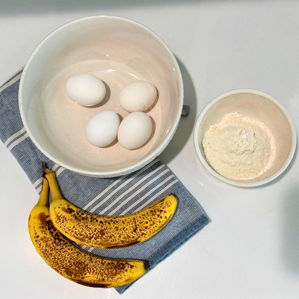 3 ingredinets for pancakes on counter: ripe bananas, eggs in a bowl and flour in a cup