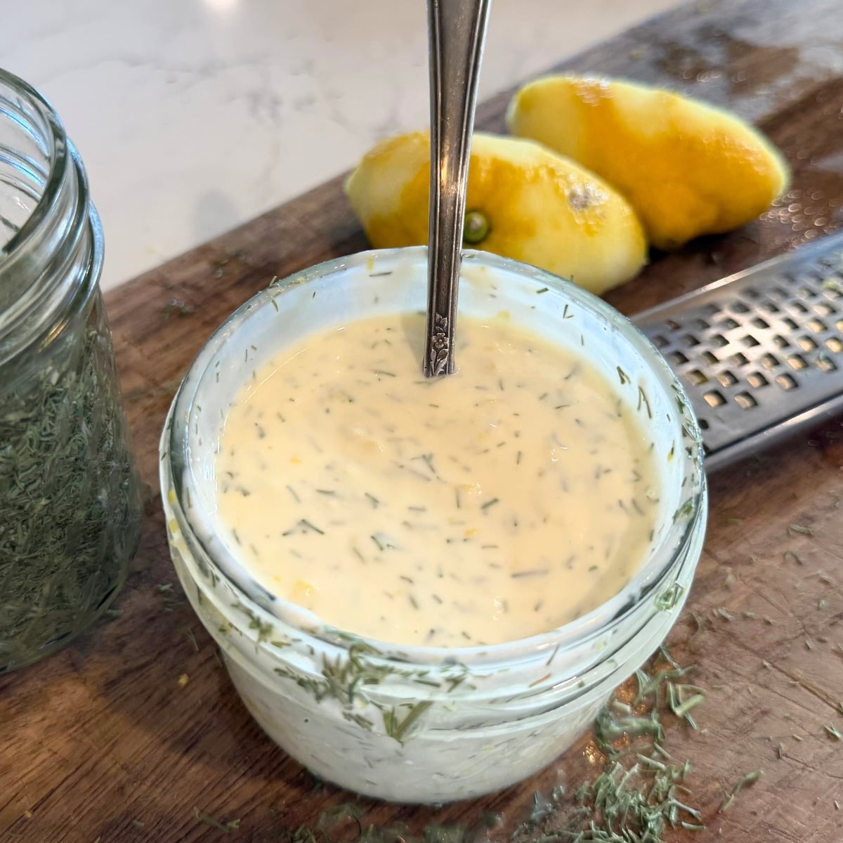 lemon dill cream sauce in a mason jar on a cutting board with dill on the cutting board and zested and juiced lemon halves.