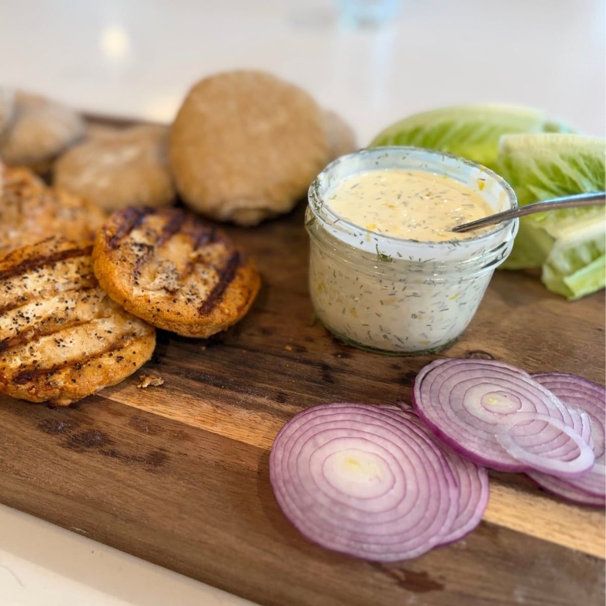 lemon dill cream sauce on a cutting board with grilled salmon patties, sourdough buns, sliced red oniona nd lettuce leaves