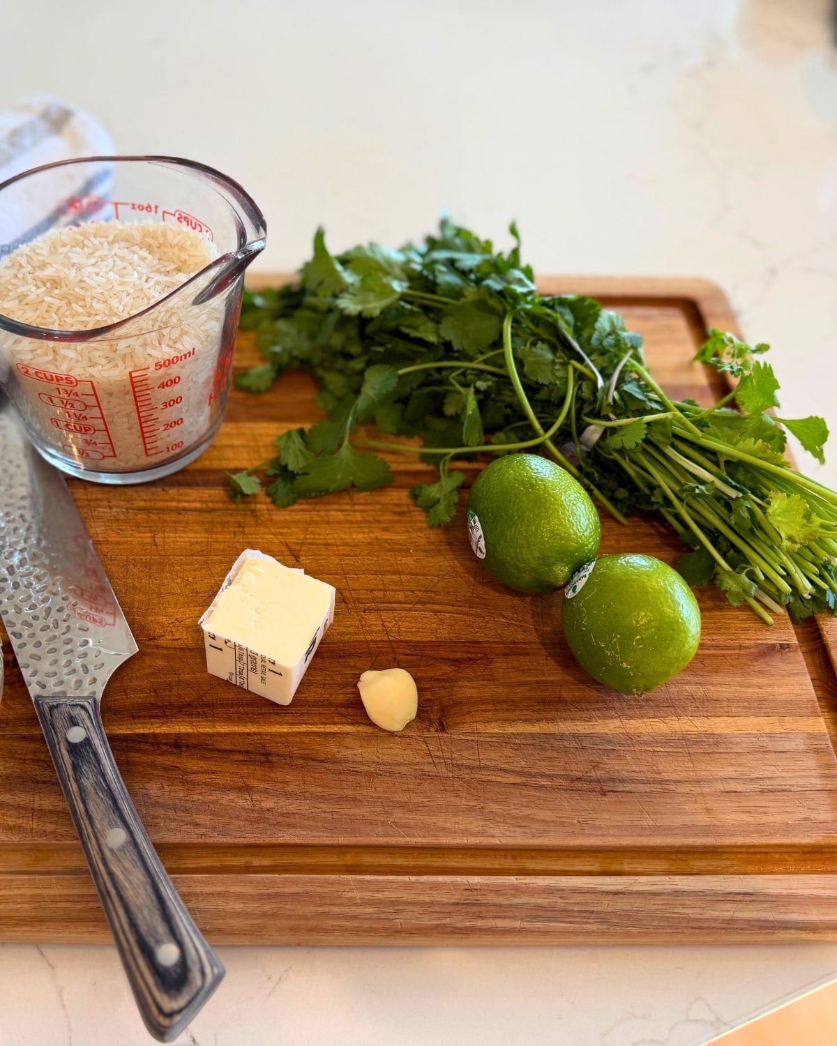 cilantro lime rice ingredients on a cutting board butter, garlic clove, jasmine rice, cilantro, and 2 limes with a chopping knife