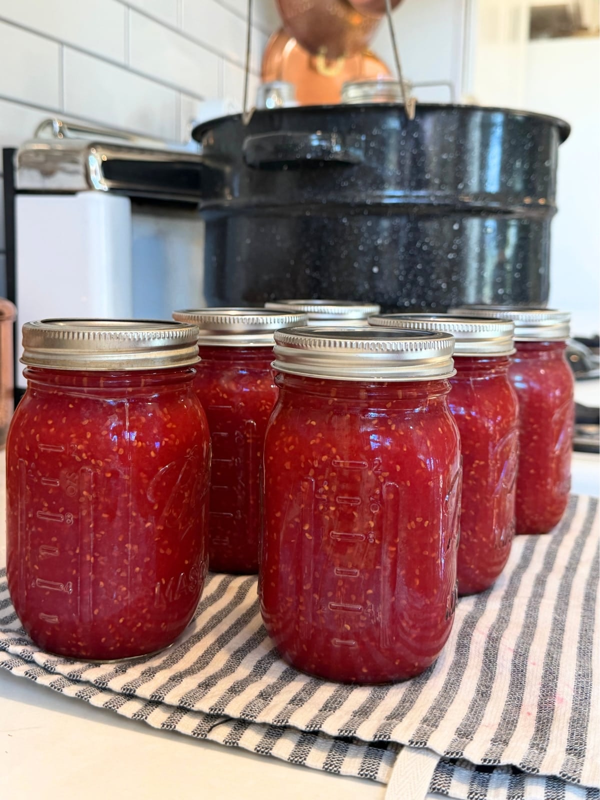 6 pint sized mason jars of hot canned raspberry jam on a kitchen towel on counter cooling with water bath canner in background
