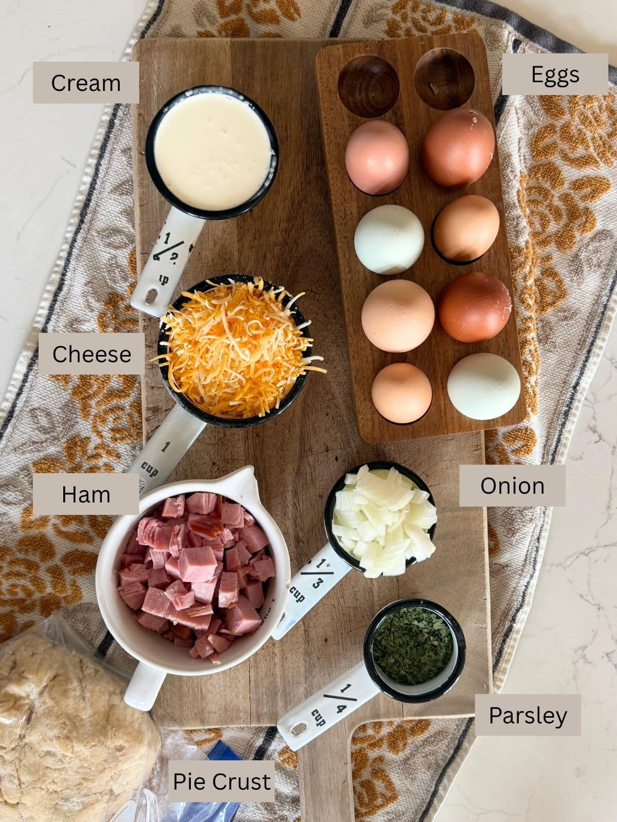 Cream, eggs, cheese, ham, onion, parsley and pie crust dough on a wooden cutting board on a towel on the counter
