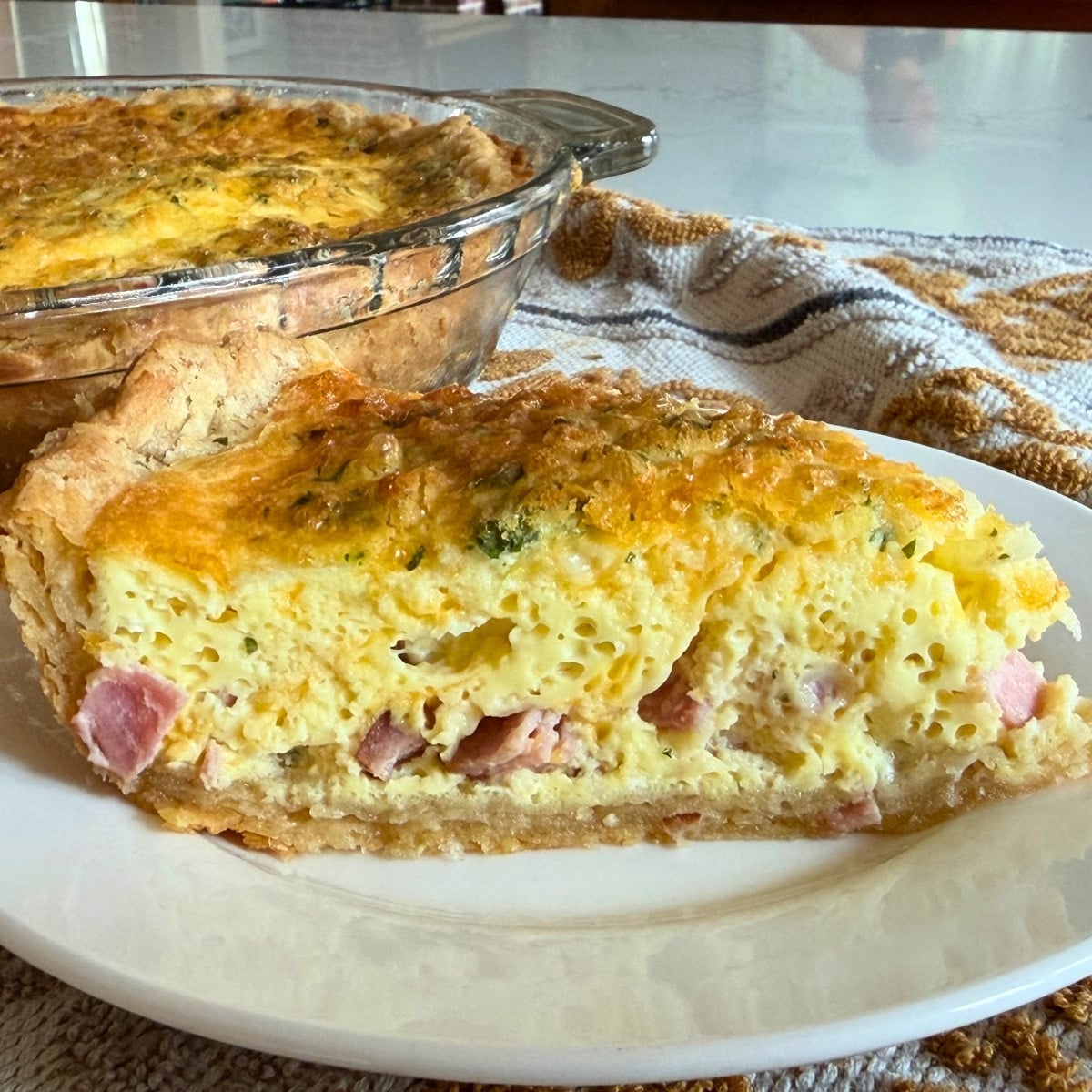 a slice of this easy quiche recipe with ham and cheese served on a white plate showing pieces of ham and parsley inside a perfectly golden brown crust and quiche with golden cheesey topping