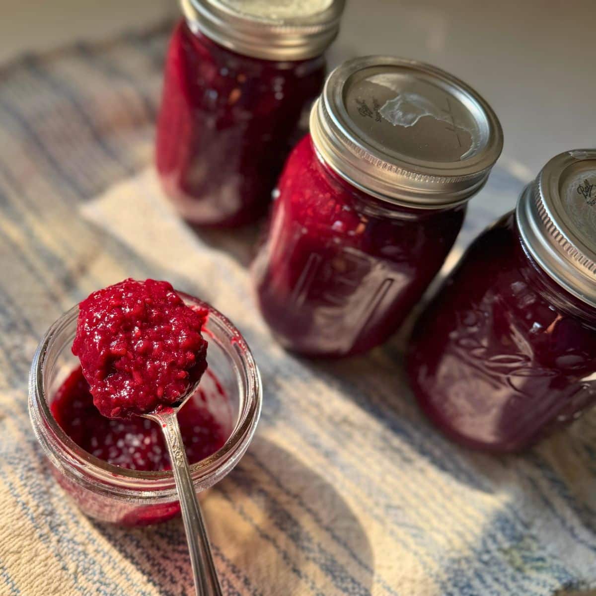 a spoonful of Honey Blackberry Jam Canning Recipe next to 3 pint sized jars of it on a kitchen towel