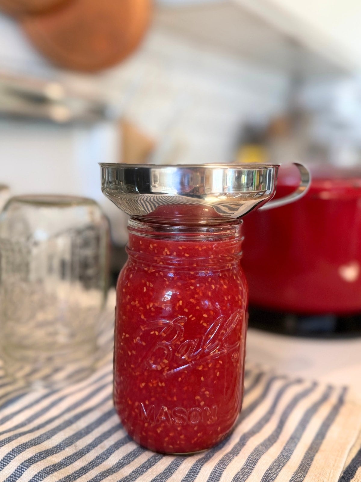 filling a pint sized mason jar with a metal funnal on top and the pot of raspberry jam in the background on the stove top