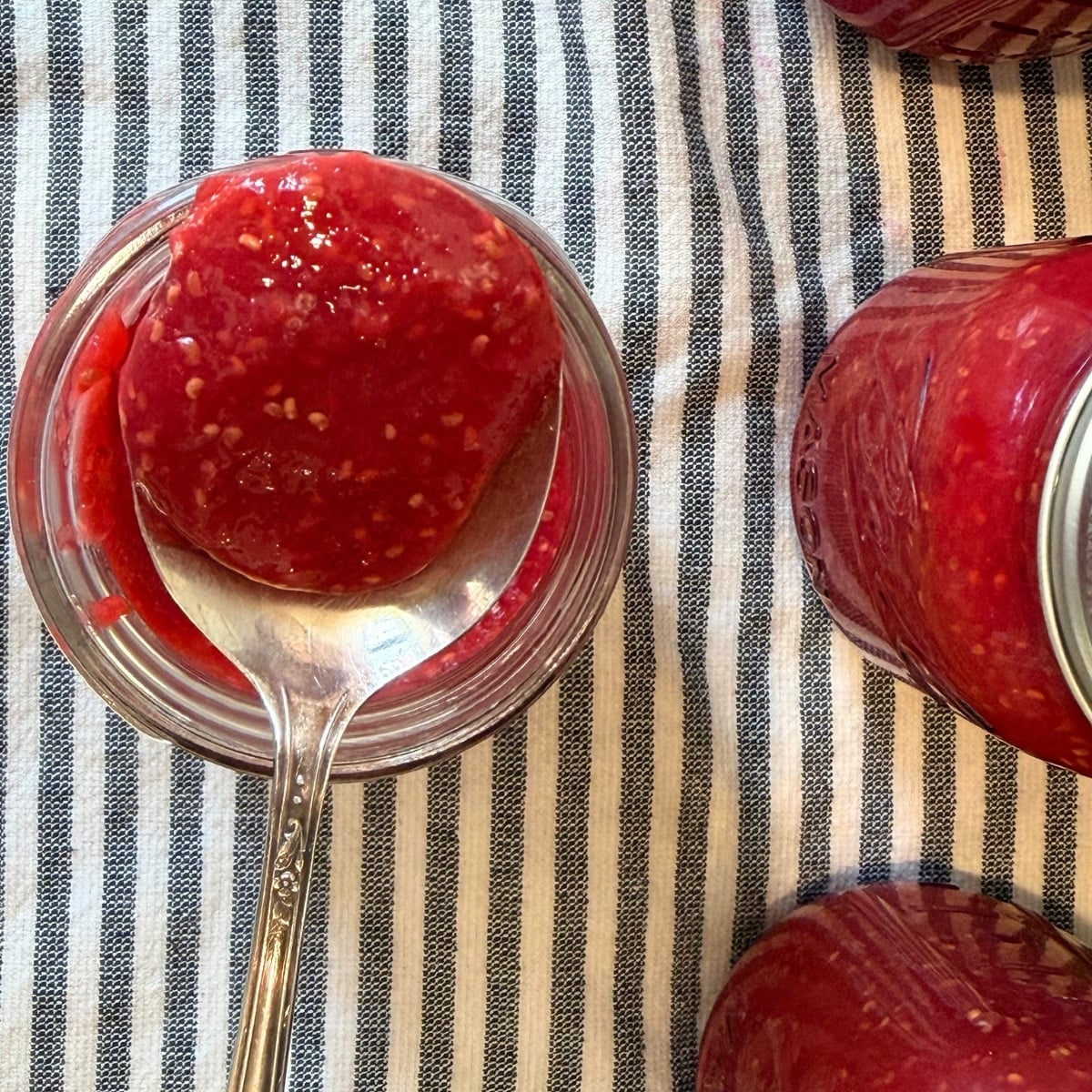 raspberry jam canning reicpe on spoon above mason jar with mason jars of raspberry jam on the side