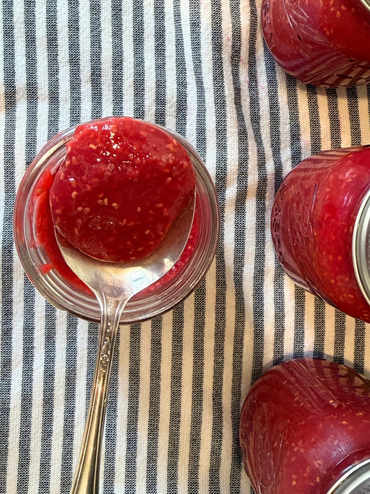 raspberry jam canning recipe on a spoon abive the pint sized mason jar beside 3 more jars of raspberry jam on a striped kitchen towel