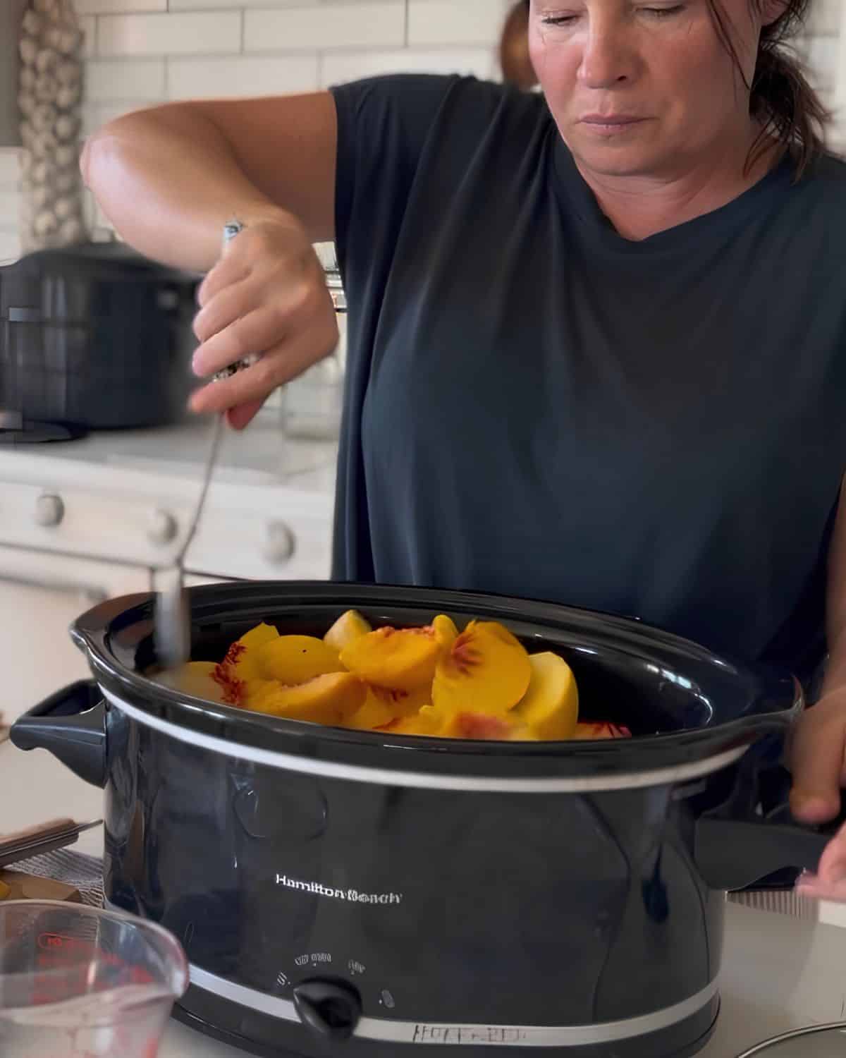 juliea from farmhouse harvest making peach butter canning recipe in the slow cooker stirring peaches in the crockpot with sugar